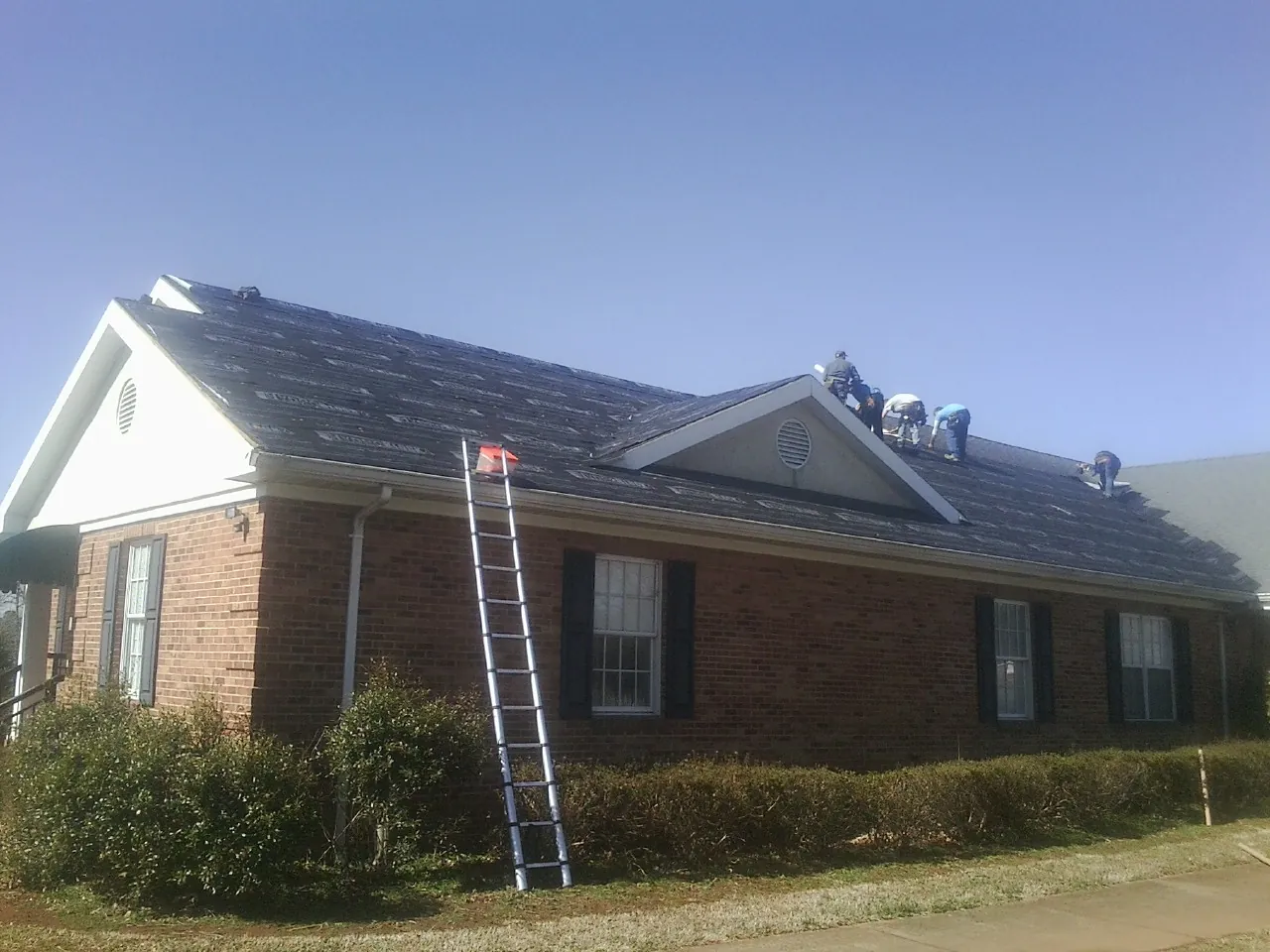 Crew tearing shingles off a brick ranch roof with a ladder set — Metal Roof Repair work by our crew in Shirley