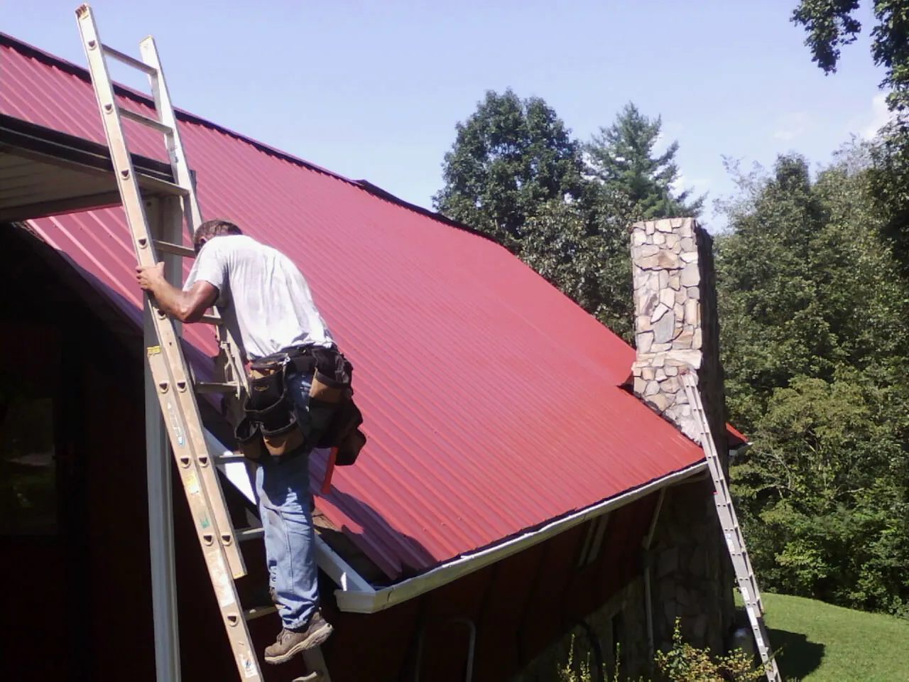 Roofer on a ladder beside a red metal roof and stone chimney — Soffit & Fascia Repair by Roofing Brothers in Shirley